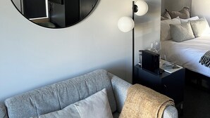 Une chambre d’hôtel moderne avec un miroir rond, un canapé gris, un lit avec une literie blanche et une table de chevet sur laquelle se trouvent des lunettes et une télécommande.