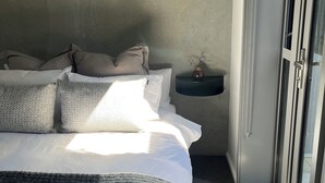 Un lit bien fait, avec une serviette pliée, une table de chevet ornée d’un vase et une fenêtre avec des stores.