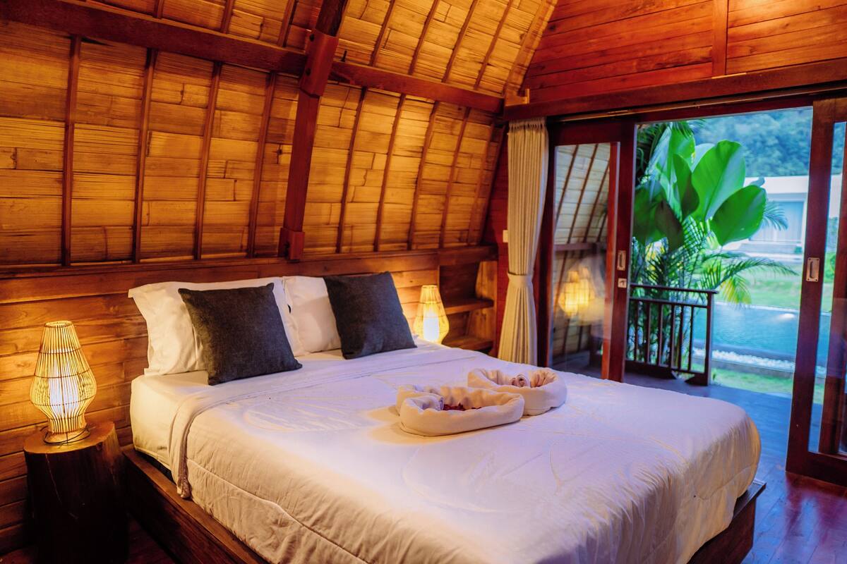 A wooden-framed bedroom with a bed, pillows, and a bedside lamp.