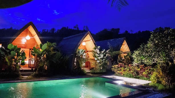 Vista nocturna de un complejo turístico con dos cabañas de madera, una piscina y exuberante vegetación tropical.