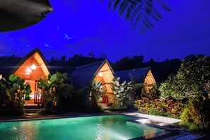A nighttime view of a resort with two wooden cabins, a swimming pool, and lush tropical vegetation. - The Caldera Villas Kintamani (Kintamani)