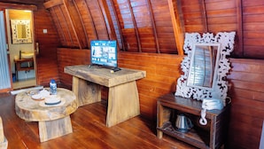 Un interior de madera con techo de madera, una mesa de madera con una computadora, una mesa redonda de madera con un sombrero y una botella, y un espejo en un estante de madera.