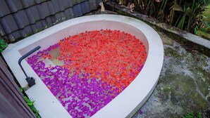 Una bañera con forma de corazón llena de bombas de baño rojas y púrpuras, rodeada de plantas y un muro de azulejos.