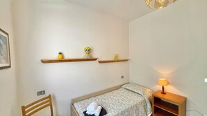Ein kleines, ordentlich eingerichtetes Schlafzimmer mit einem Einzelbett, einem Sessel, einem Nachttisch und zwei Wandregalen mit Dekoration.