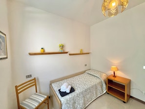 Ein kleines, ordentlich eingerichtetes Schlafzimmer mit einem Einzelbett, einem Sessel, einem Nachttisch und zwei Wandregalen mit Dekoration.