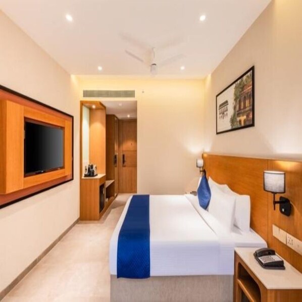 Une chambre d’hôtel dotée d’un grand téléviseur à écran plat fixé au mur, d’un lit avec une literie blanche agrémentée d’une touche de bleu, d’une table de chevet avec une lampe et d’un tableau encadré au mur.
