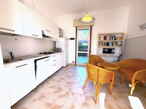 A kitchen with white cabinets, a gas stove, a refrigerator, and a dining area with wicker chairs and a table. - VILLA DUNE NORTH 289 - TYPE A2 6+0 - B02899 (Bibione Pineda)