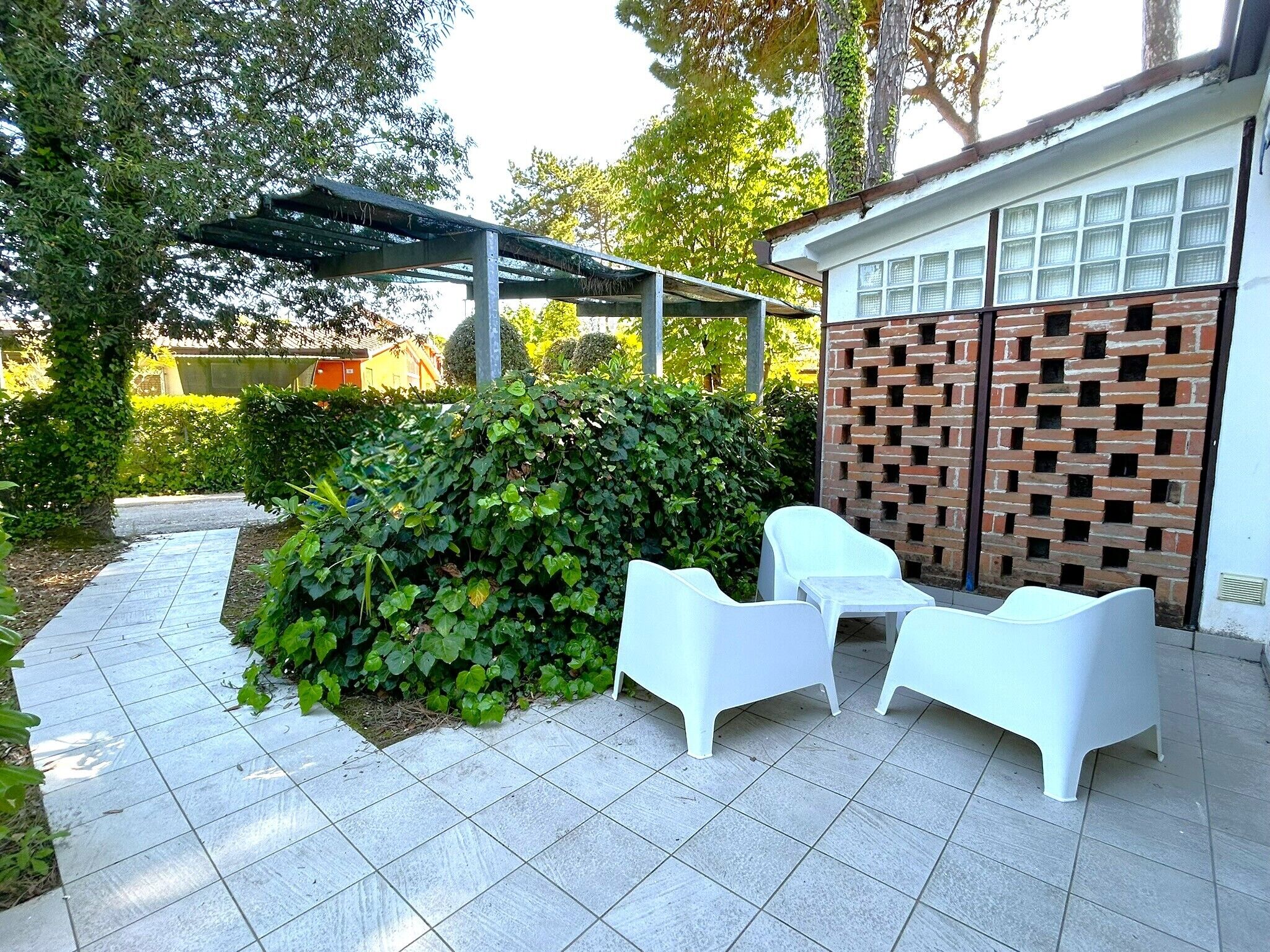 A patio with tiled flooring, white outdoor furniture, a brick wall, and a green hedge.