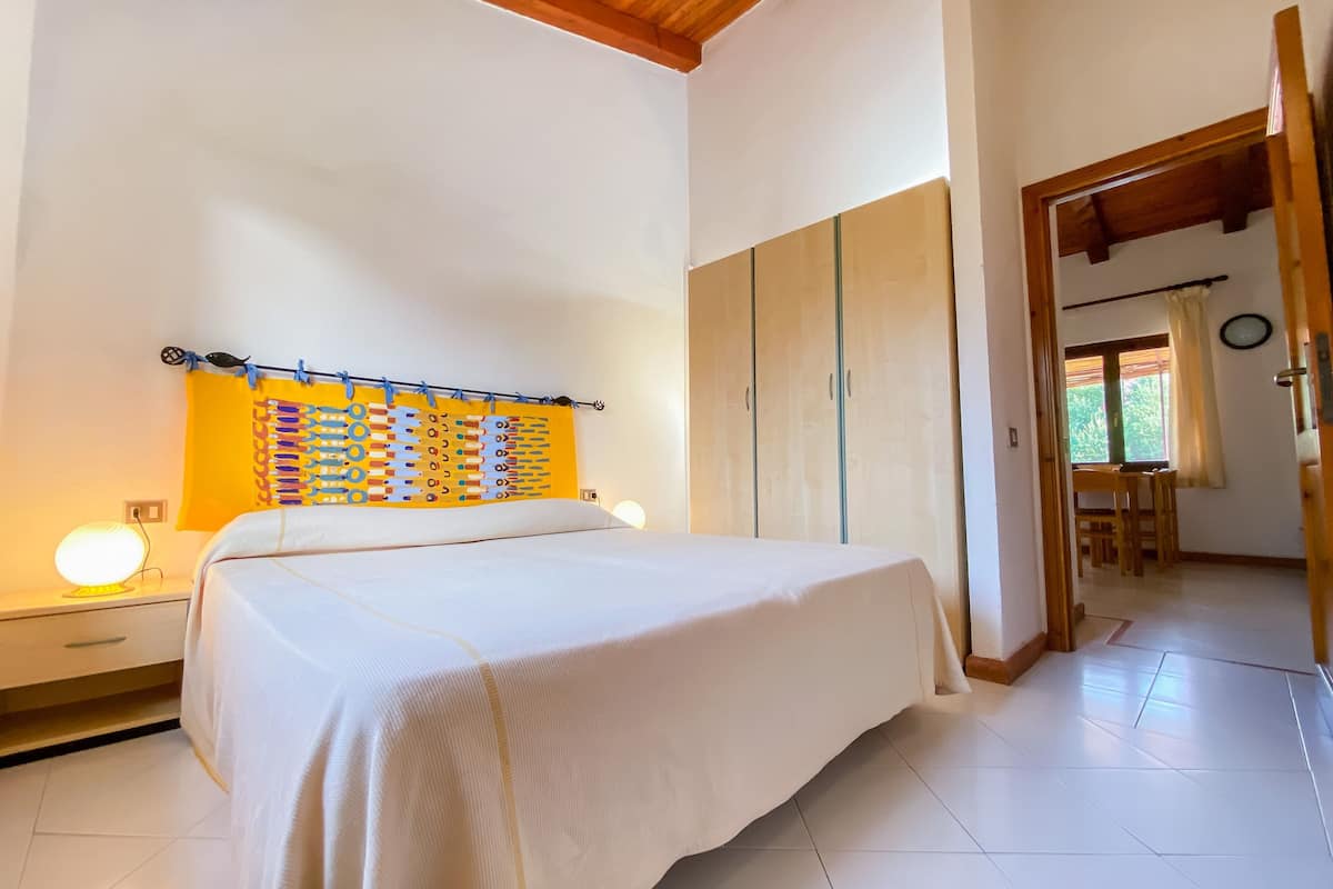 A bedroom with a bed, a nightstand, a wall-mounted lamp, a wardrobe, and a dining area with chairs and a table.