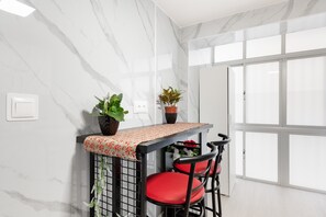 Eine Küchenzeile mit Marmorfliesenspiegel, ein Fenster mit Jalousien, ein Tisch mit gemustertem Tischläufer und zwei rote Stühle.
