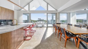 A modern kitchen with a central island, a dining area with a long table and chairs, and large windows offering a view of the outdoors. - Magic Beach Robe - Luxury Beachfront Escape (Robe)