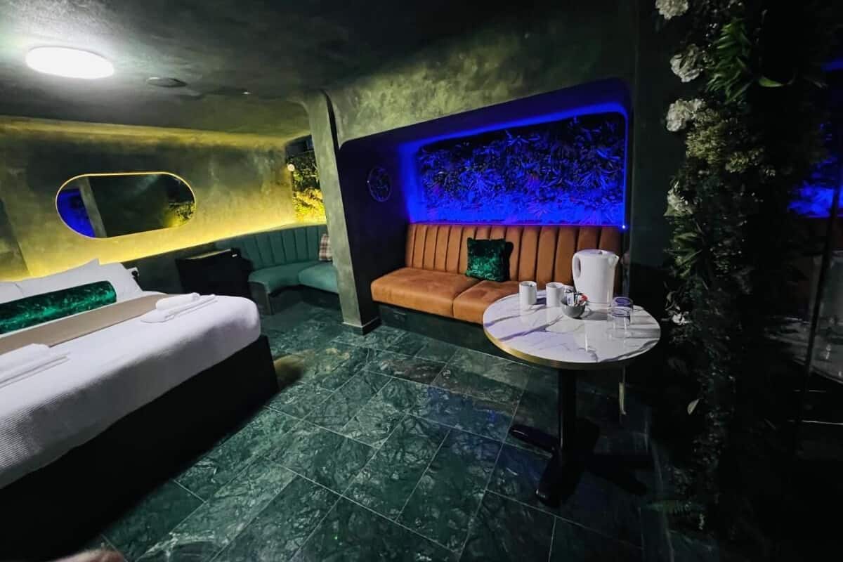 A dimly lit room with a bed, a sofa, and a small table with a vase and a bowl.