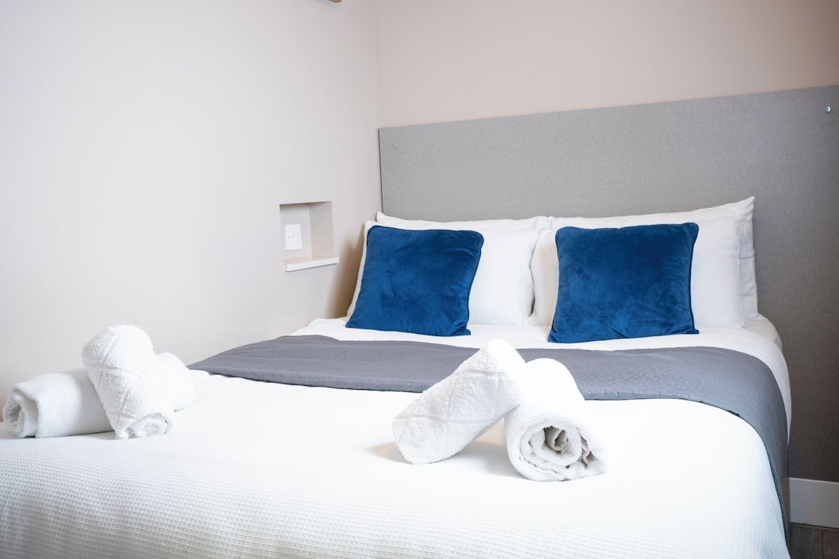 A neatly made bed with white and blue bedding, two rolled towels, and a gray accent pillow.