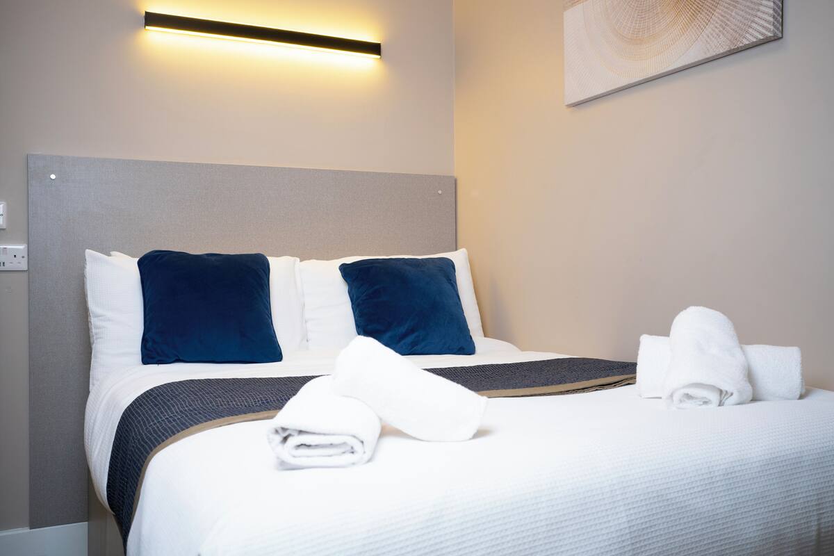 A neatly made bed with white and dark blue pillows, a folded white towel, and a rolled-up towel on a textured white bedspread.