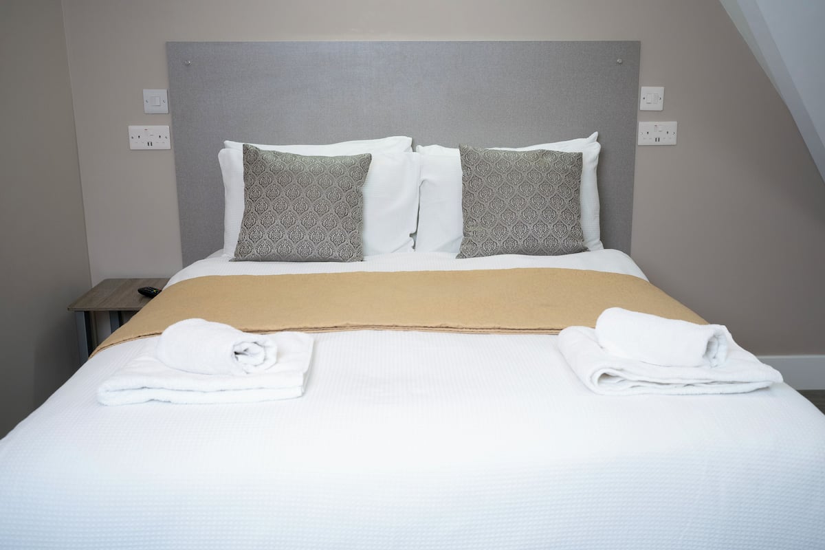 A neatly made bed with a beige bedspread, two patterned pillows, and two folded white towels on either side.