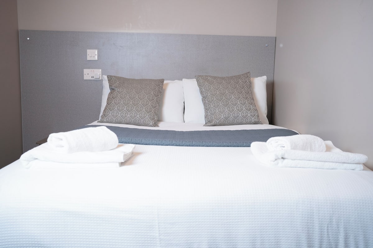 A neatly made bed with white linens and gray patterned pillows, a gray headboard, and a white wall with a power outlet.