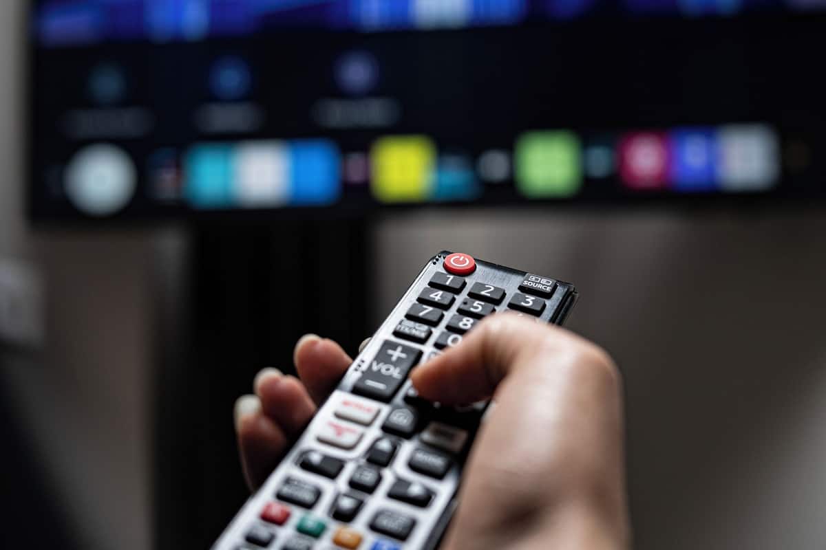 A hand holding a remote control in front of a television displaying a news broadcast.