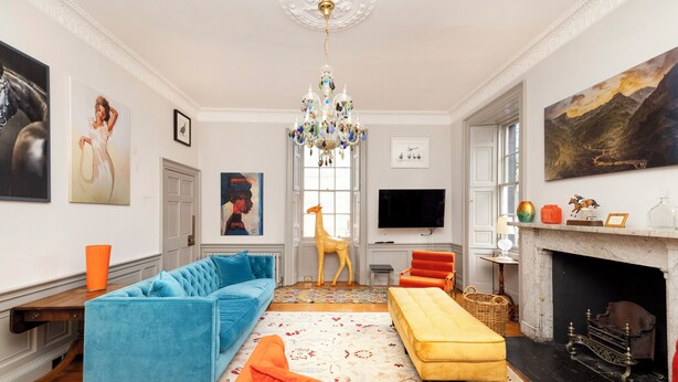 Ein Wohnzimmer mit einem blauen Sofa, einem gelben Hocker, einem Kamin und einer Kristallleuchtre.