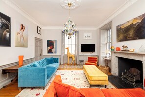 A living room with a blue sofa, a yellow ottoman, a fireplace, and a chandelier. - Luxury Family Flat in Edinburgh New Town (Edinburgh)
