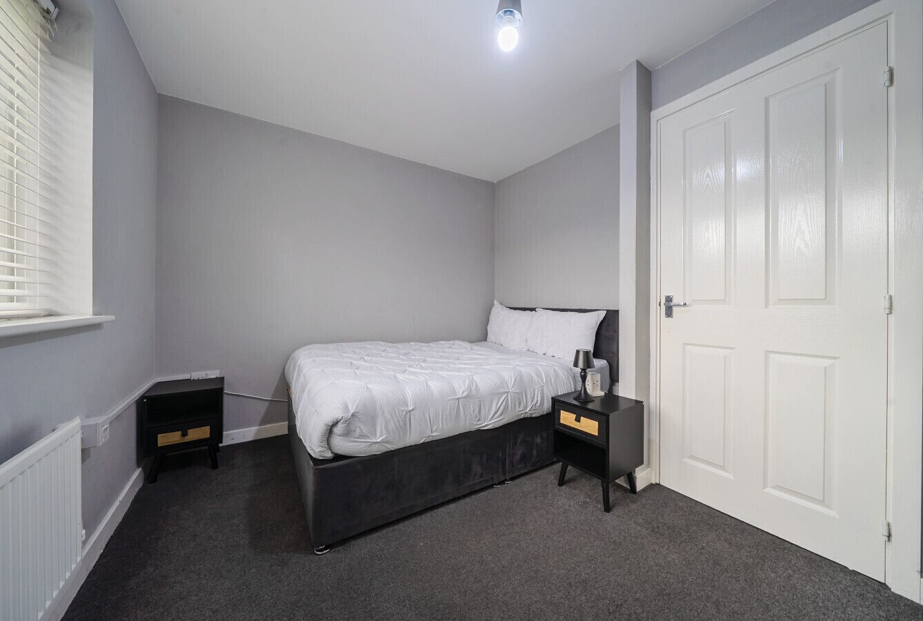 A bedroom with a bed, two nightstands, a white door, and a window with blinds.