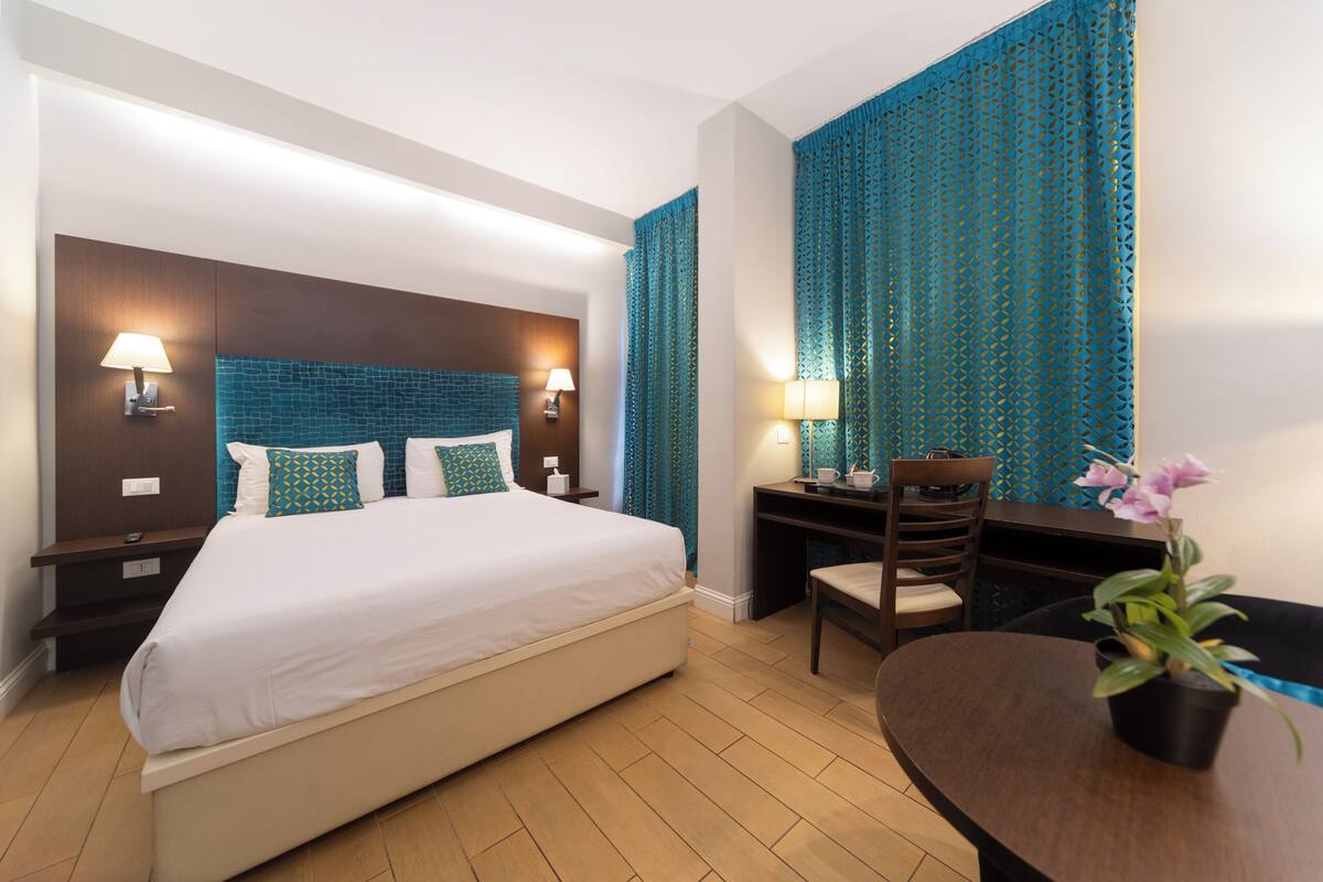 A hotel room with a large bed, two bedside tables with lamps, a desk with a chair, and a potted plant.