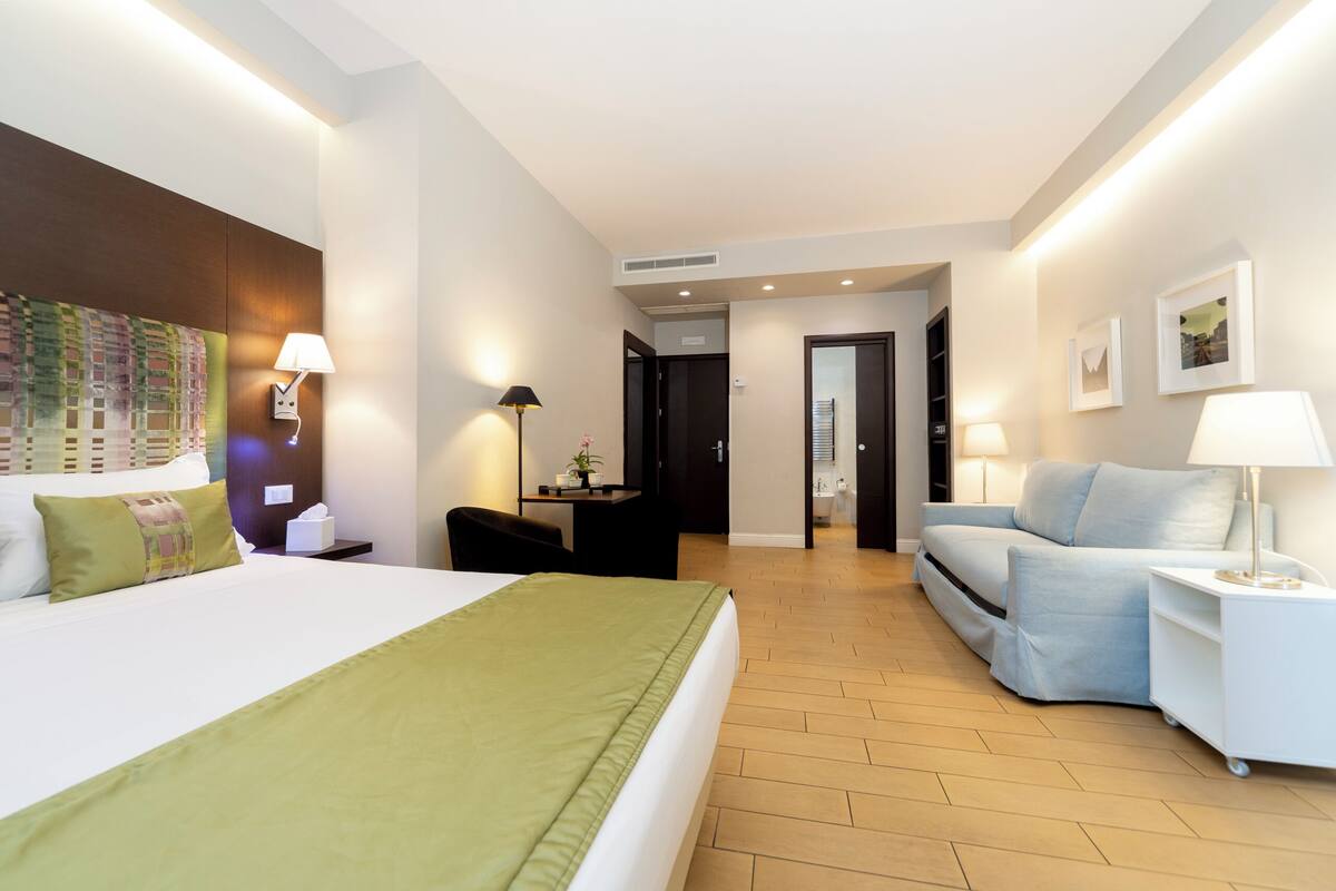 A modern hotel room with a large bed, a sofa, a desk, and a chair. There are wall-mounted lamps and a door leading to another room.