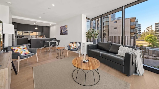 A modern apartment with a living room, dining area, and kitchen. It features large windows, a black leather sofa, and a wooden coffee table. - Bright Unit with Secure Parking and Garden Access (Reid)