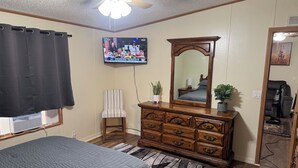 A bedroom with a bed, dresser, mirror, ceiling fan, TV, and a window with curtains.