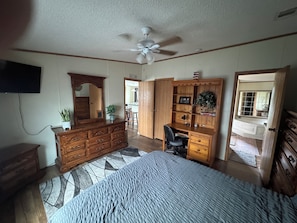 Un dormitorio con cama, cómoda, escritorio, silla, ventilador de techo y televisor montado en la pared.