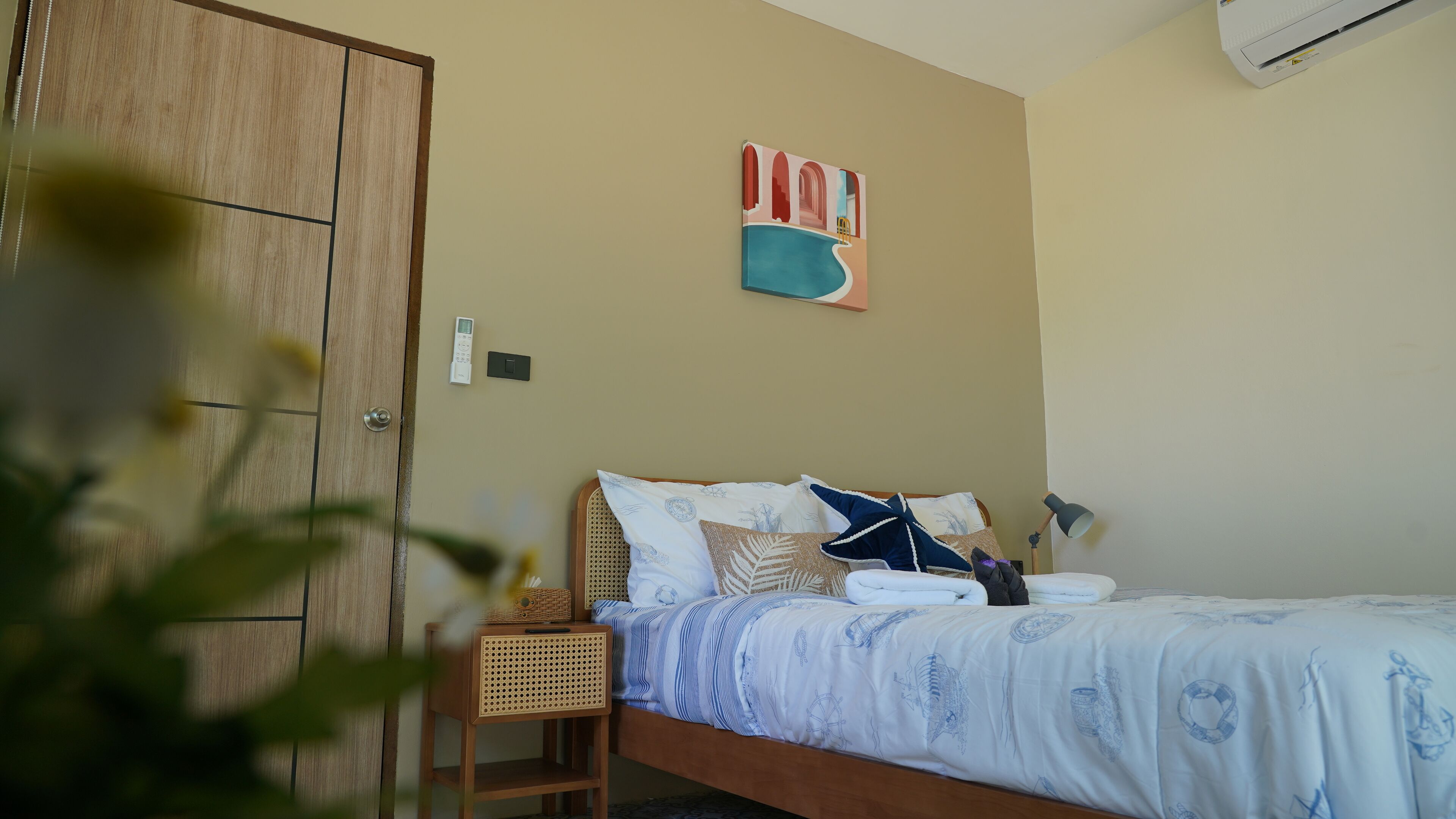 A bedroom with a bed, a bedside table, a wall-mounted air conditioner, and a painting.