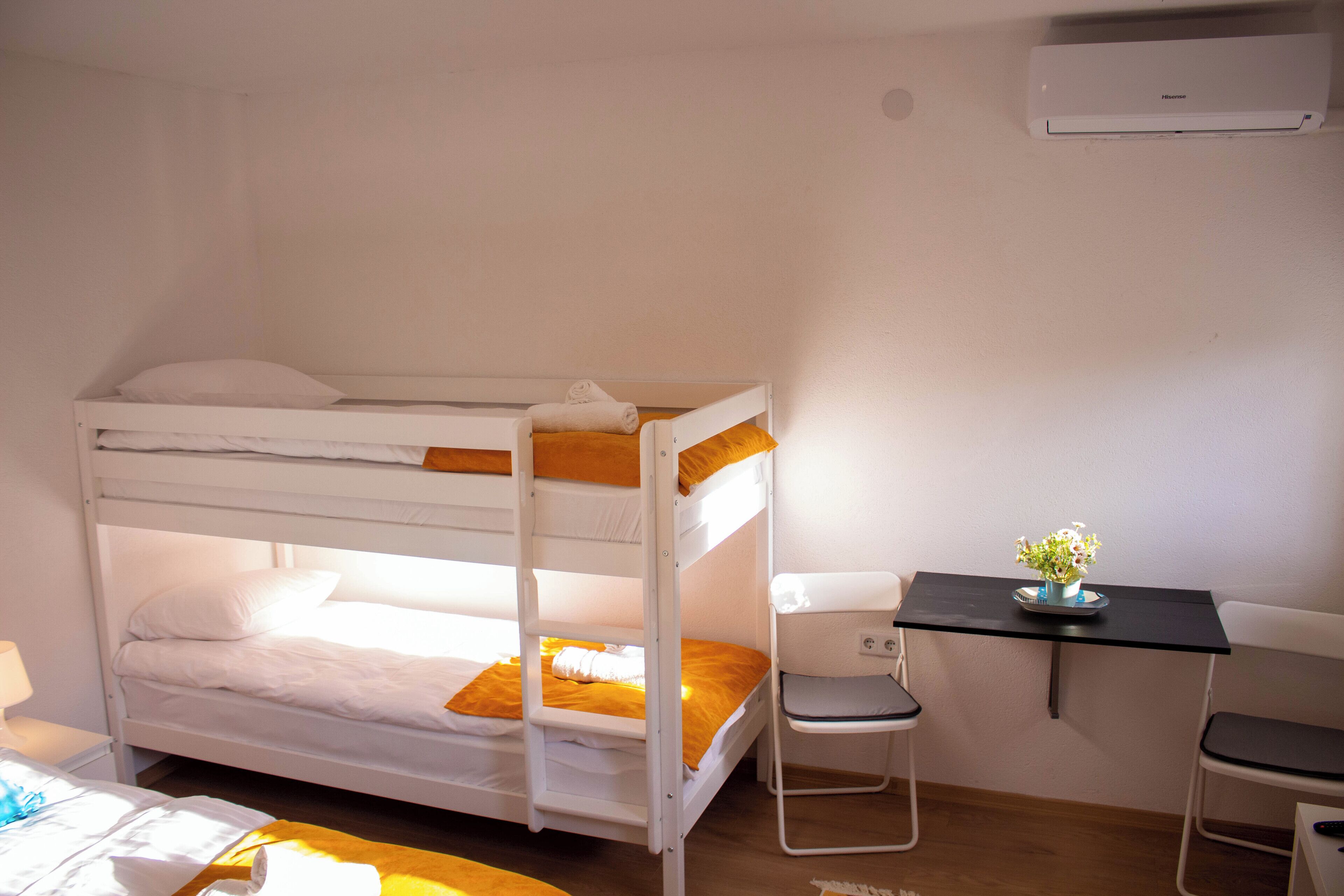 Una cama litera con escritorio y silla, una mesita con una planta y un equipo de aire acondicionado en la pared.
