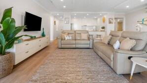 Una sala de estar moderna con un sofá beige, un televisor de pantalla plana y una cocina al fondo.