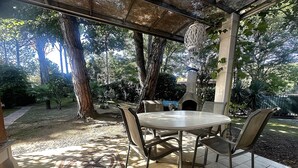 An outdoor patio with a round table and chairs, surrounded by trees and a stone fireplace. - VILLA DUNE NORTH SD4 - TYPE B2 4+0 - B00043 (Bibione Pineda)