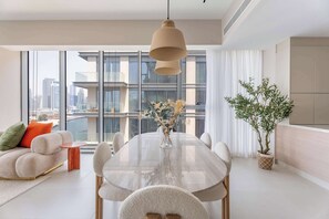 A modern living room with a large dining table, comfortable seating, a large window with a city view, and a potted plant. - Elegant 3BR + Office | Canal View | Pool & Gym Access (Al Wasl)