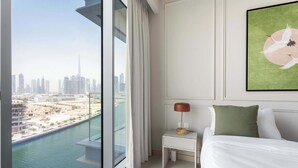 Ein Hotelzimmer mit großem Fenster und Blick auf die Stadt, einem ordentlich bezogenen Bett, einem kleinen Nachttisch mit Lampe und einem gerahmten Kunstwerk an der Wand.
