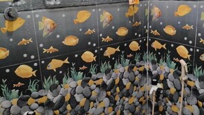 A bathroom with a tiled wall depicting an underwater scene with fish and dolphins, a shower area with a red bucket, and a towel rack. - VRS INN (Walajapet)