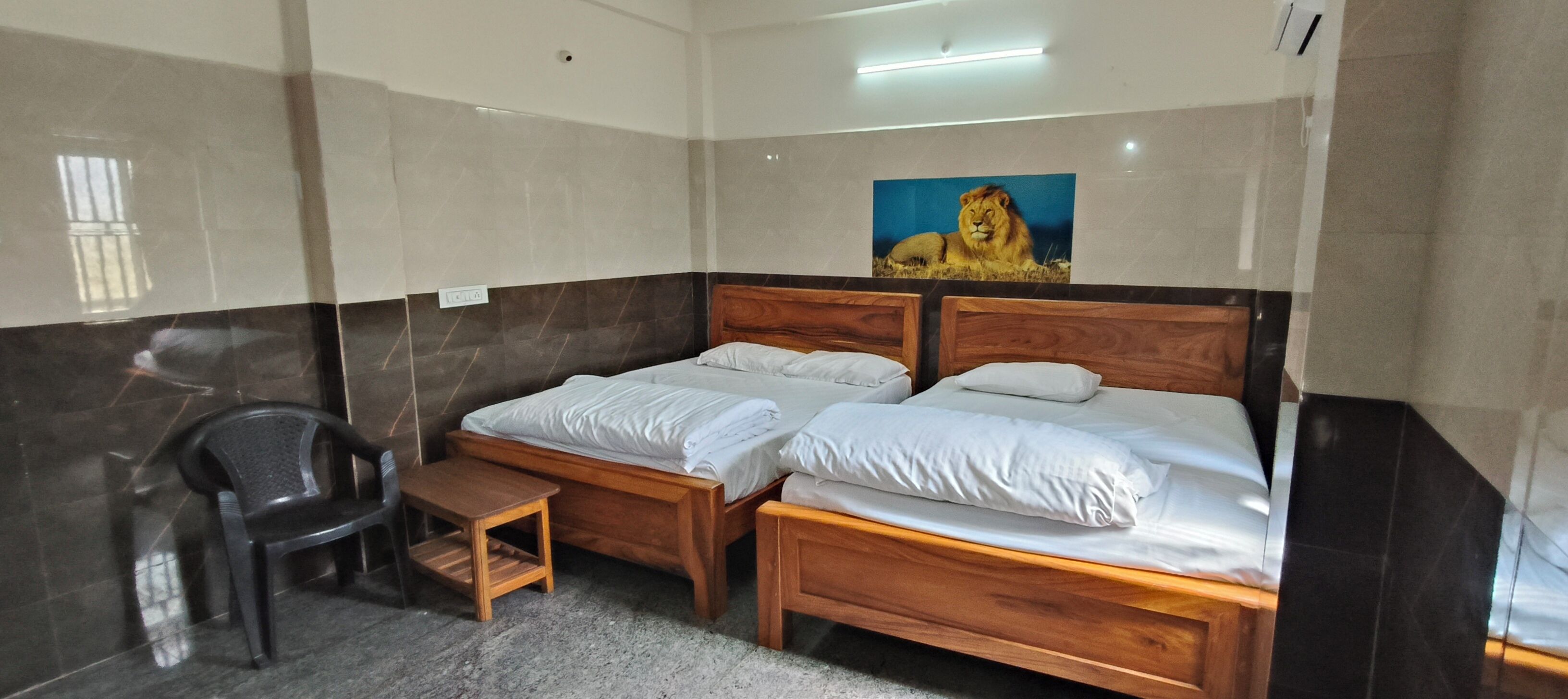 A hotel room with two wooden beds, a small wooden table, a plastic chair, and a large painting of a lion on the wall.