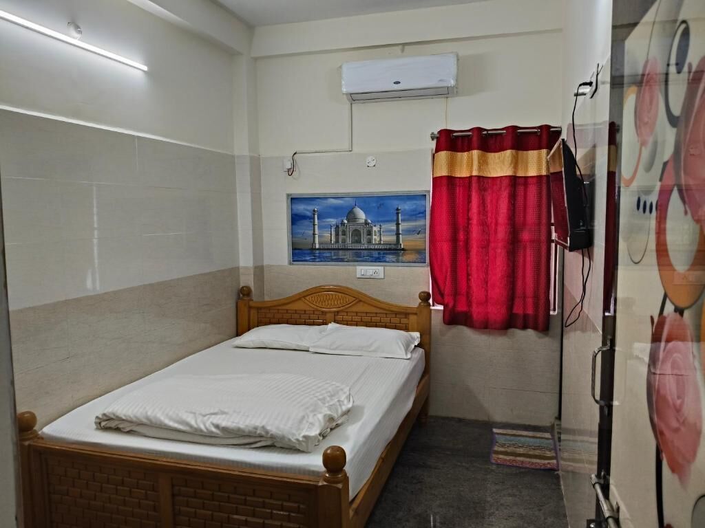Un lit simple avec une tête de lit en bois, un climatiseur fixé au mur, un tableau représentant le Taj Mahal et un rideau rouge.