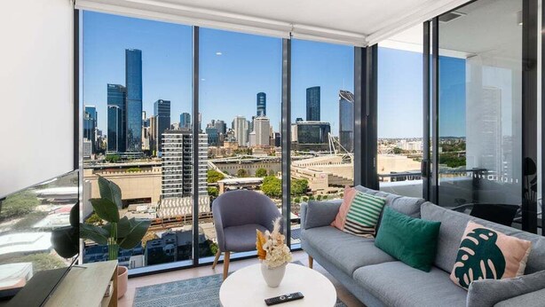 A modern living room with a large window offering a city view, a grey sofa with decorative pillows, a round coffee table with a vase and remote control, and a plant on the side table. - Top Location Convenient City View Modern Living (Brisbane)