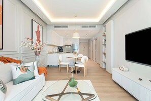 A modern living room with a sofa, coffee table, and a flat-screen TV. There is a dining area with a table and chairs, and a kitchen in the background. - Luxury Renovated 1BR w Full Sea View (Dubai Marina)