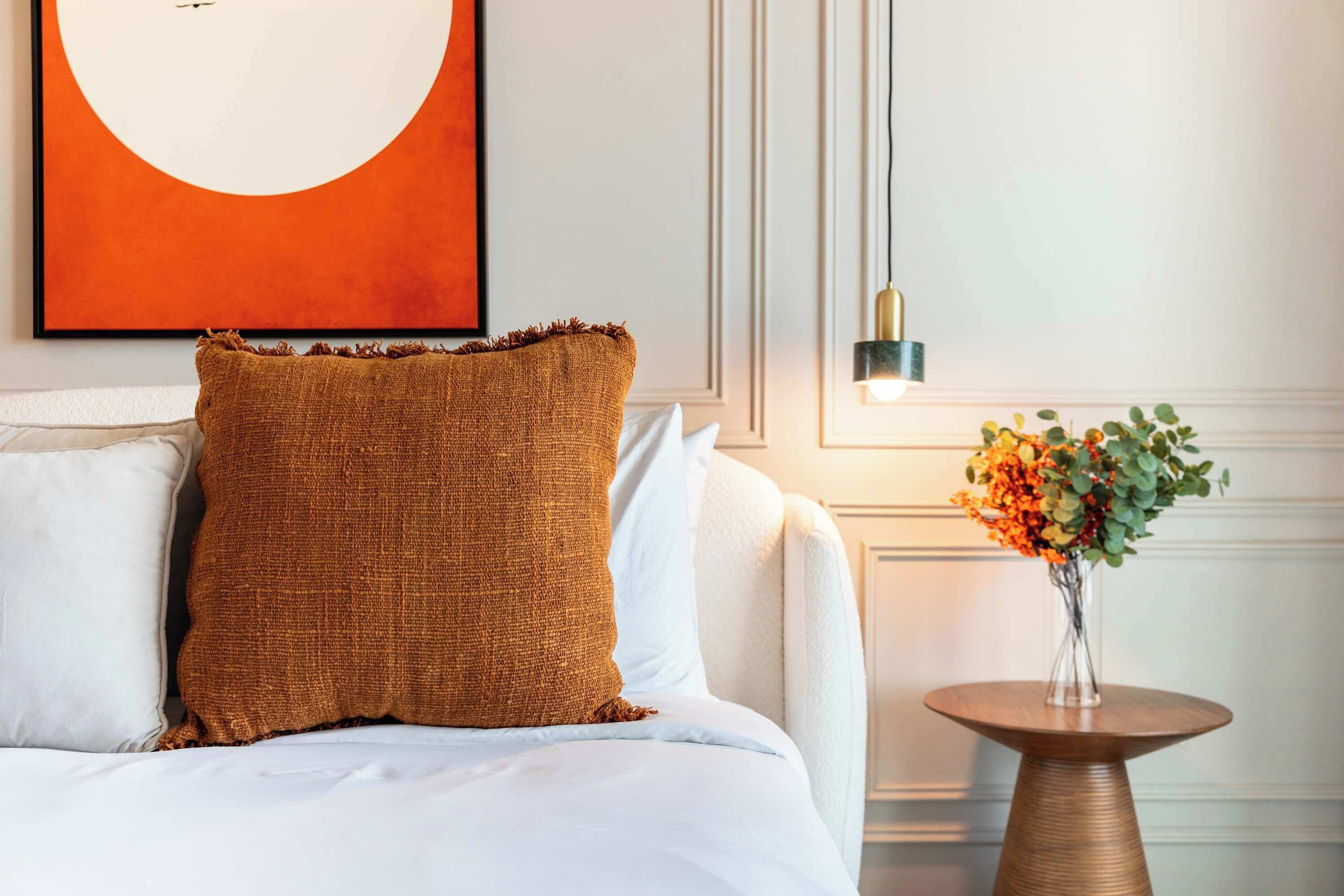 A neatly made bed with a textured brown pillow and a white pillow, a round wooden side table with a vase of flowers, and a wall-mounted light fixture.