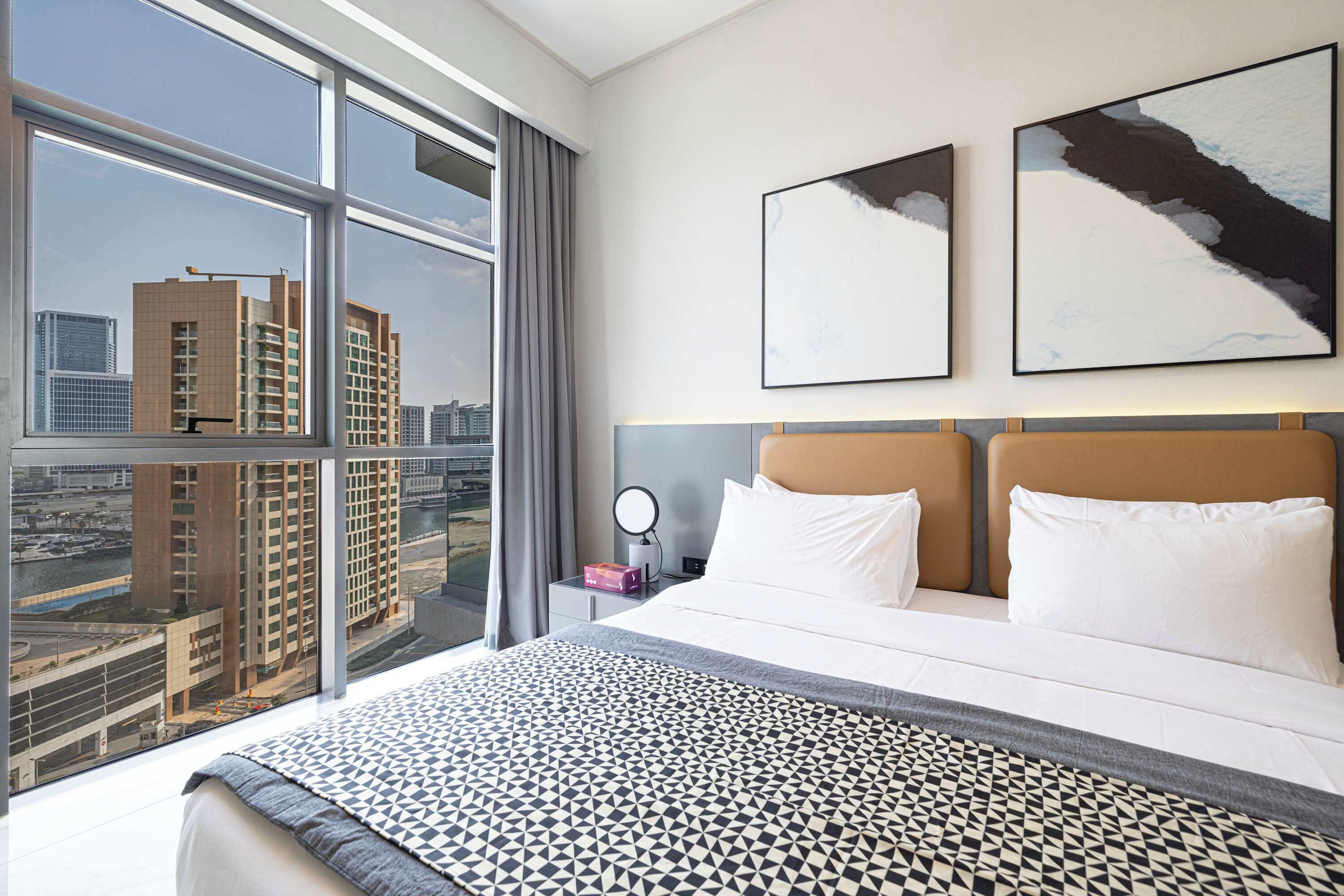 A hotel room with a large bed, two framed artworks, a nightstand with a mirror, and a view of a cityscape through large windows.