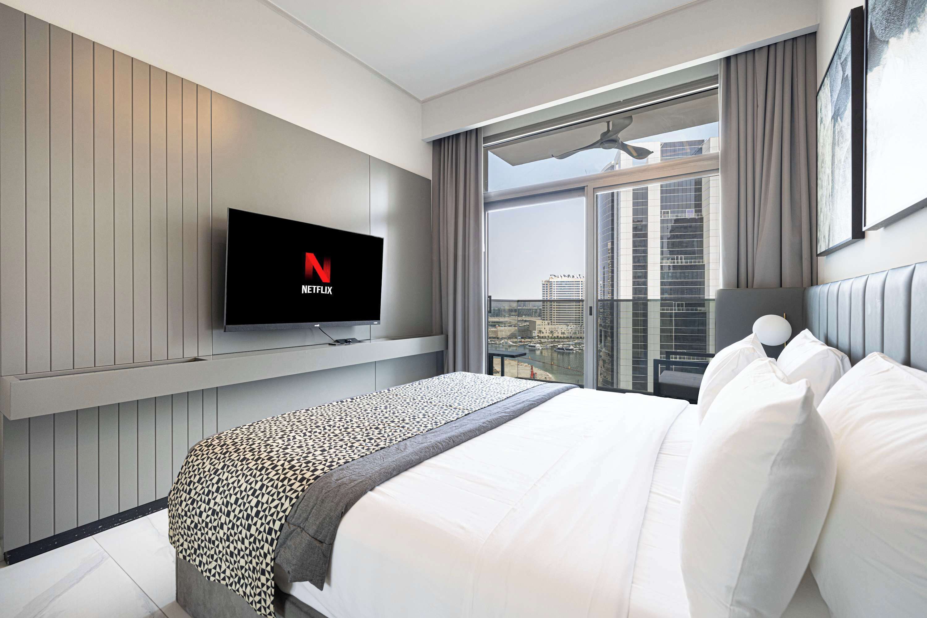 Une chambre d’hôtel moderne avec un grand lit, un téléviseur fixé au mur affichant le logo de Netflix, une vue sur la ville à travers de grandes fenêtres et un balcon donnant sur le paysage urbain.