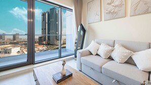 Un salón moderno con un sofá gris, una mesa de centro con un libro y vistas a un paisaje urbano a través de grandes ventanales.