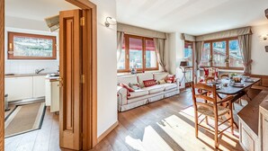 Eine moderne Küche mit Holzschränken, ein Essbereich mit Holztisch und -stühlen sowie eine gemütliche Sitzecke mit Sofa und Kissen.