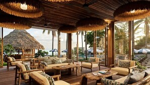 Terraza de madera con techo de paja, asientos, piscina y palmeras.