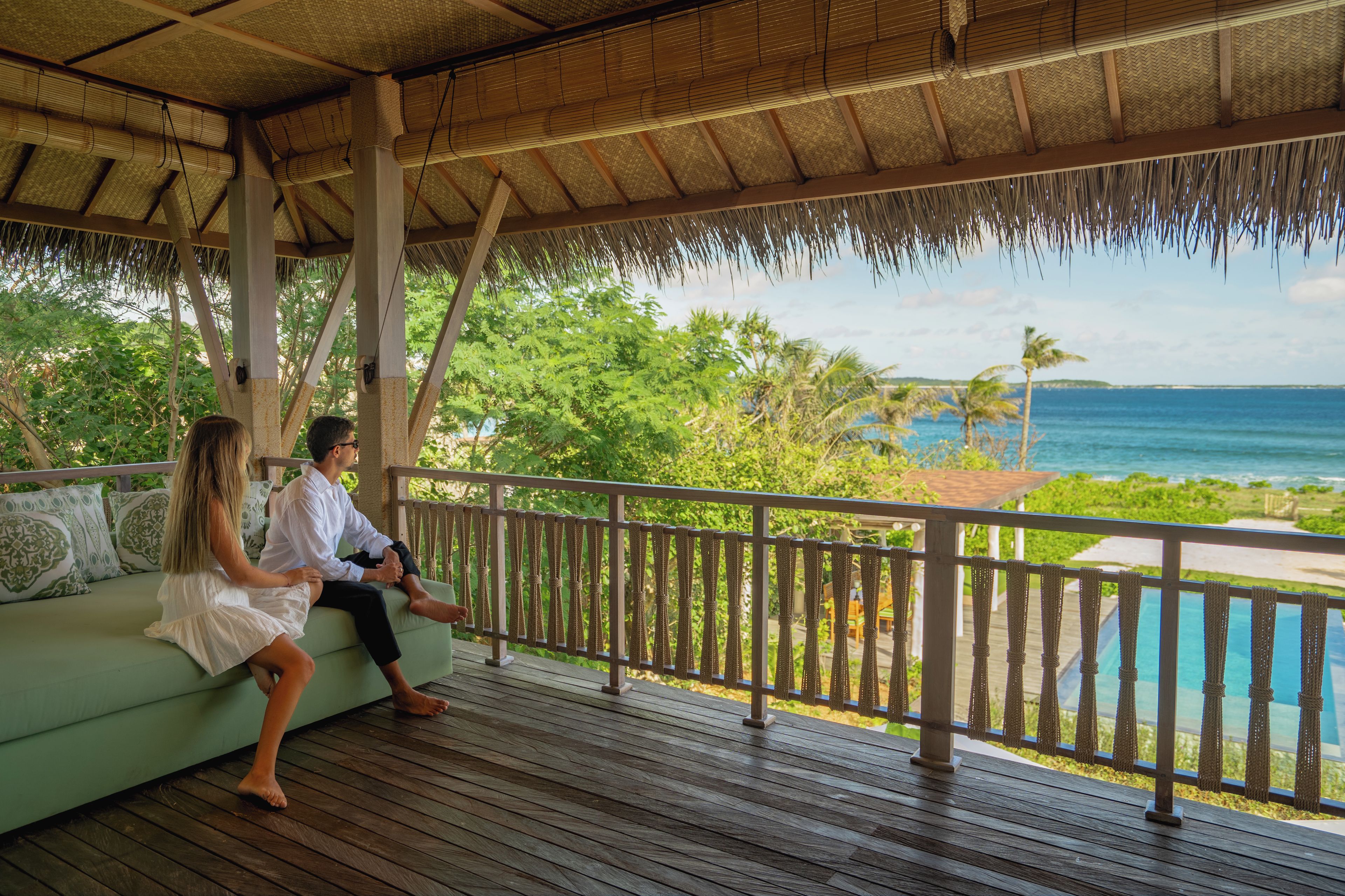 A wooden deck with a thatched roof, overlooking a pool and ocean view.