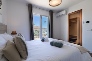A hotel room with a bed, a lamp, a window with curtains, an air conditioning unit, and a wooden wardrobe. - Villa Playa Esperanza by Rentallorca (Playa de Muro)