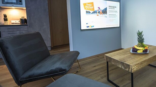 A modern living room with a flat-screen TV mounted on the wall, a wooden coffee table with fruit, and a black sofa. - Apart Inntalerhof (Toesens)