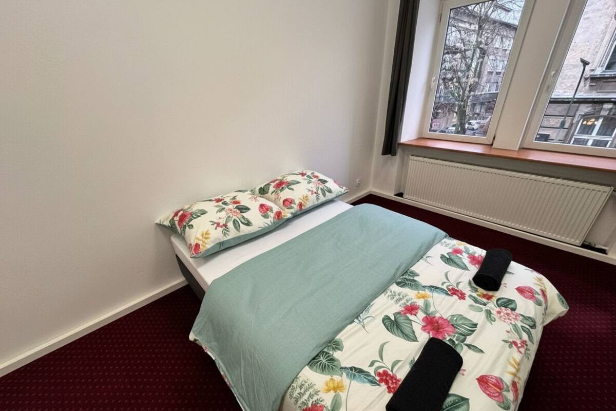 A bedroom with a bed, floral bedding, a window with a view of buildings, and a radiator.